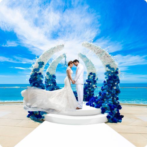 A couple in formal wedding attire stands under a large blue and white floral arch overlooking the ocean.