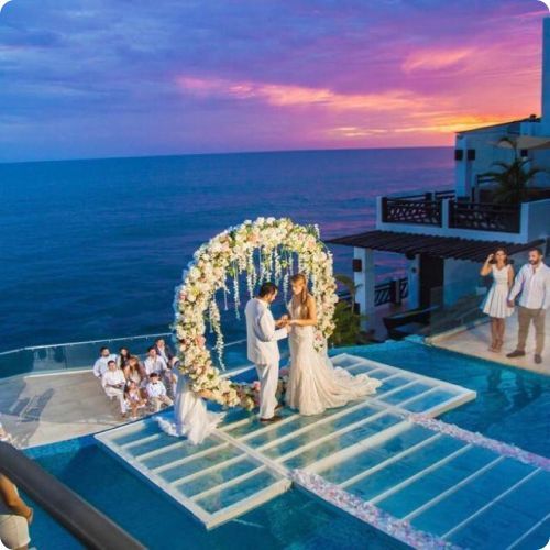 A wedding ceremony takes place under a floral arch on a glass stage over an infinity pool at sunset by the ocean.