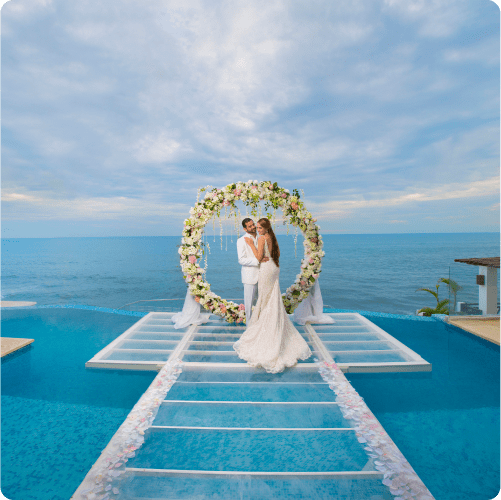 A couple stands embracing under a circular floral arch on a glass walkway extending over a turquoise swimming pool.