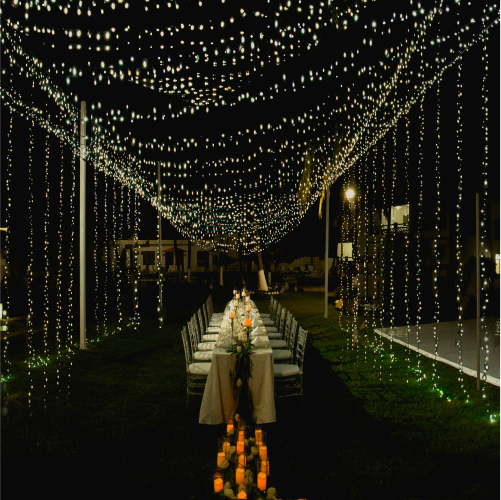 An outdoor dining table set for an event under a canopy of hanging string lights at night, with candles on the ground.