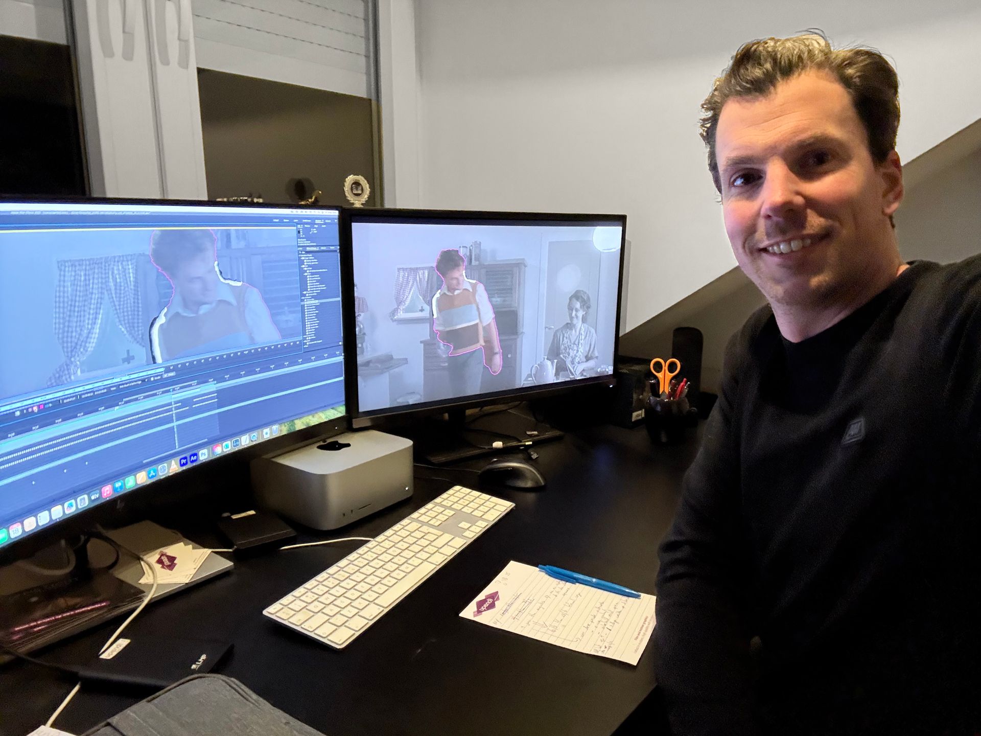 Een persoon lacht naar de camera en zit aan een bureau met twee computermonitoren waarop videobewerkingssoftware wordt weergegeven.
