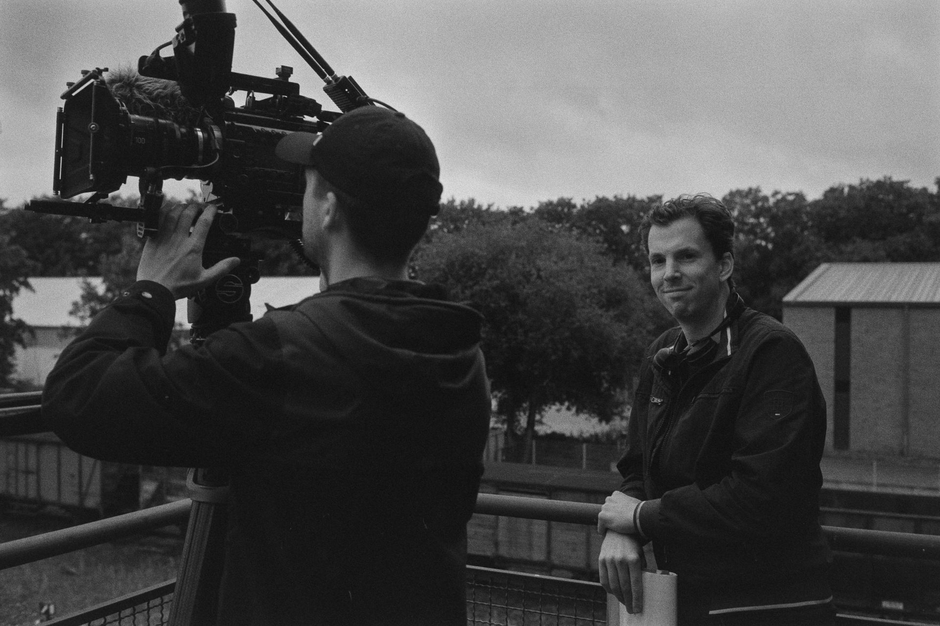 Een crewlid bedient een filmcamera op een statief, terwijl een andere persoon in de buurt staat en naar de camera kijkt.