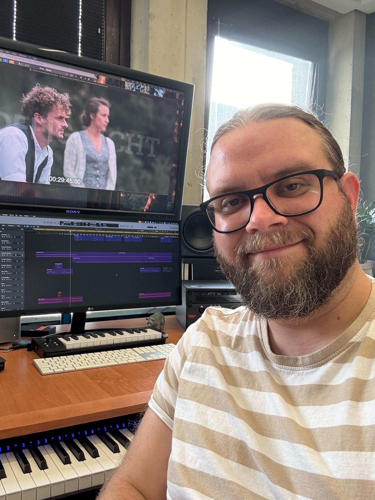 Een glimlachend persoon met een baard en een bril zit voor een computerscherm waarop videobewerkingssoftware te zien is.