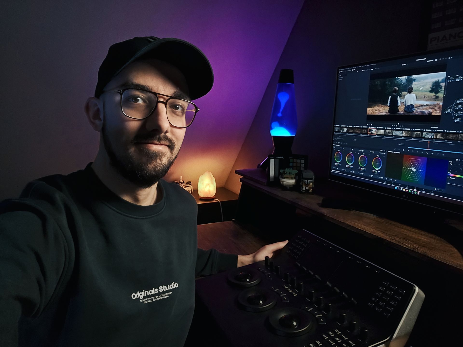 Een persoon met een bril en een pet bewerkt een video op een computer, in het schemerige, paars verlichte licht van een lavalamp en een zoutlamp.