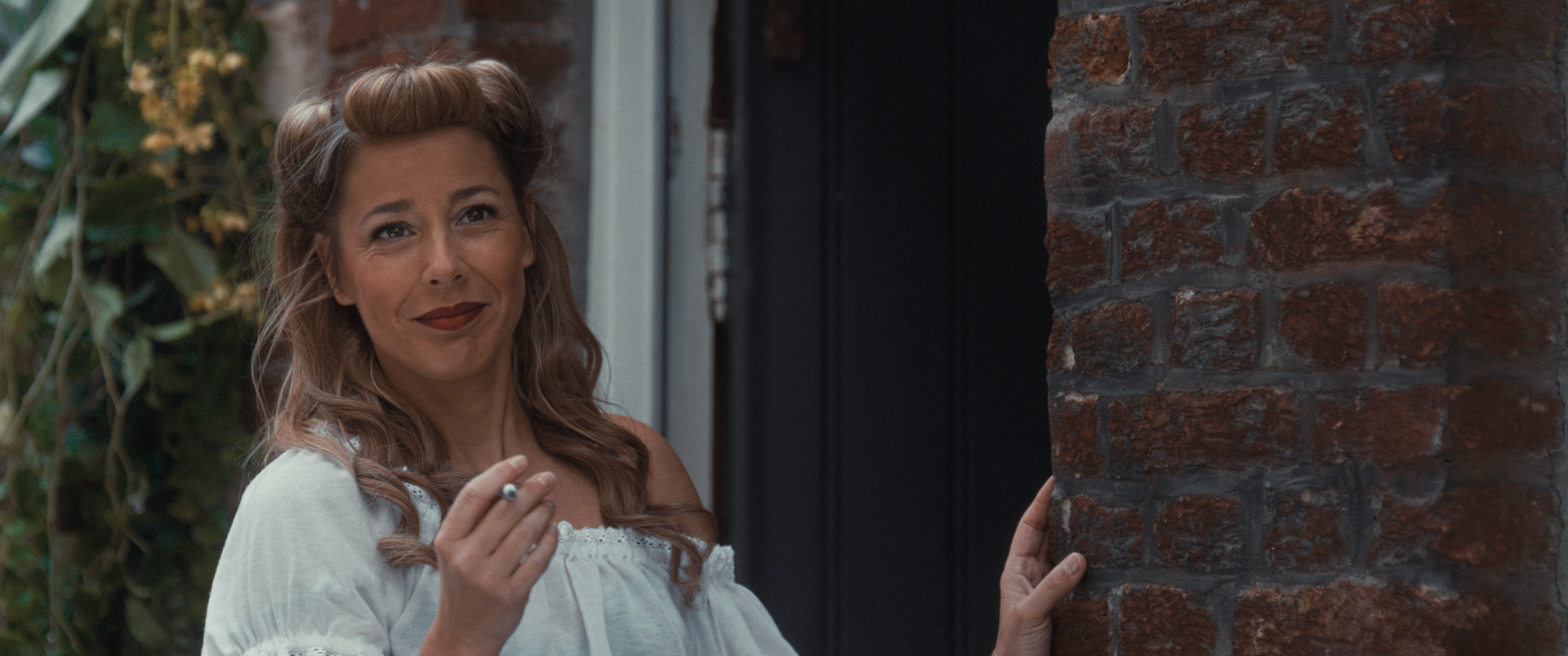 Een vrouw met golvend haar, opgestoken in victory rolls, staat met een sigaret in haar hand voor een rode bakstenen muur.