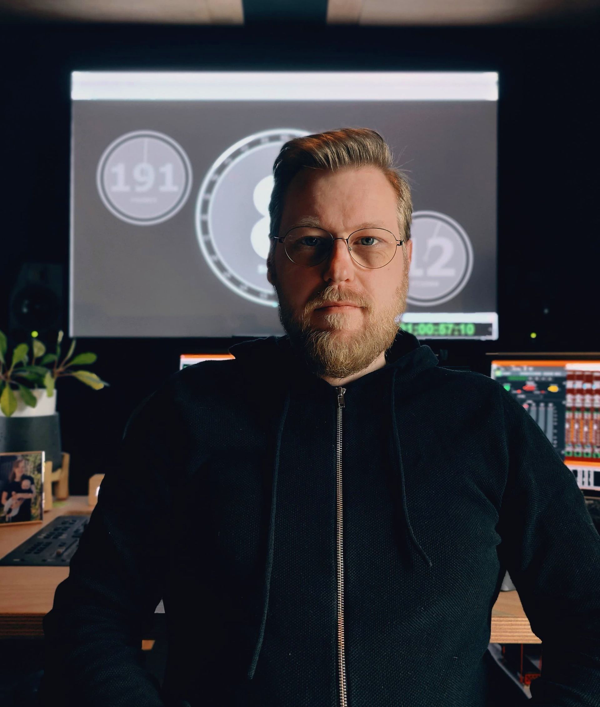 Een bebaarde persoon met een bril en een donkere hoodie met rits zit aan een bureau voor een monitor met een digitaal scherm.