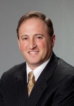 Professional headshot of a smiling man in a dark pinstripe suit and gold tie against a gray background