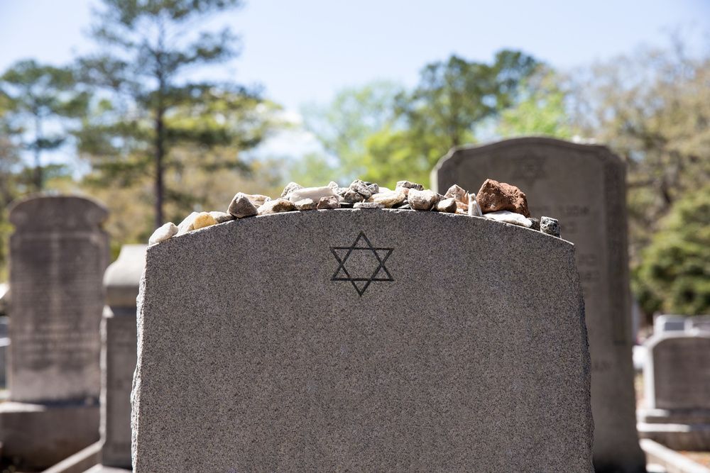 Gravestone with a Star of David in a cemetery, topped with small stones, with blurred headstones in the background