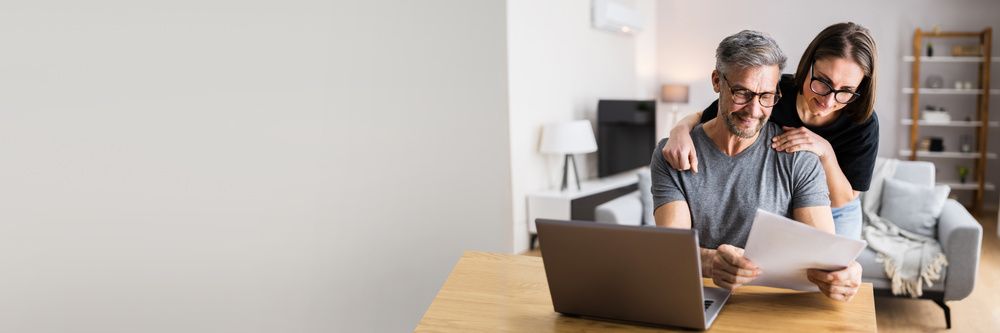 Two people review a document beside a laptop in a bright living room, with a dog resting on the couch.