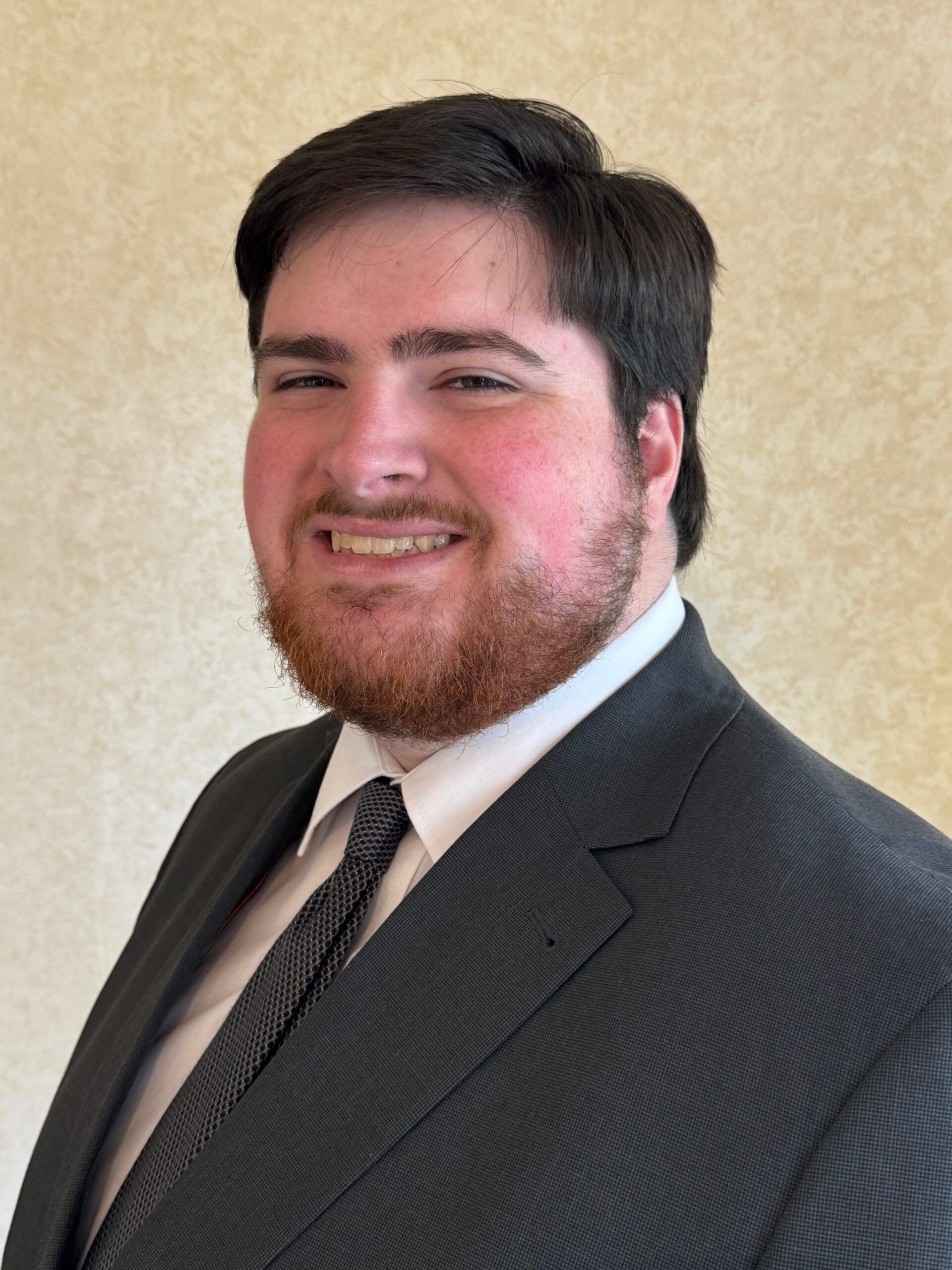 Smiling man in a dark suit and tie posing against a beige wall
