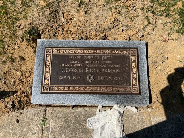 Bronze memorial plaque set in a stone base on dry ground, with flowers nearby