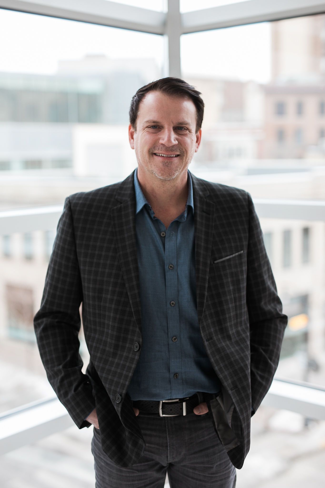 A man in a dark plaid blazer and blue button-down shirt smiling while standing in front of a bright office window.