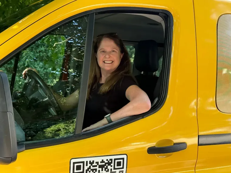Smiling woman seated in the cab of a yellow truck, visible through the open window.