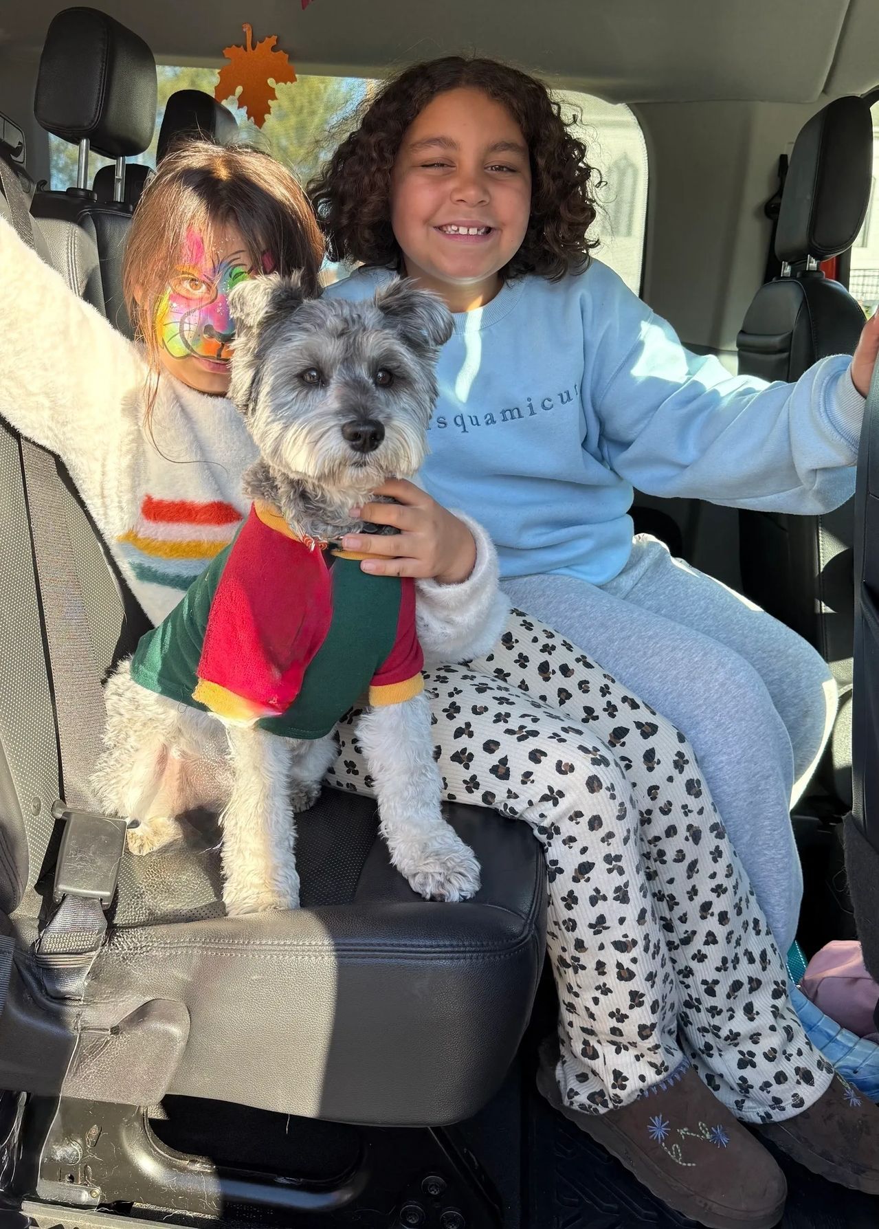 Smiling child in a car seat holding a small dog wearing a colorful sweater