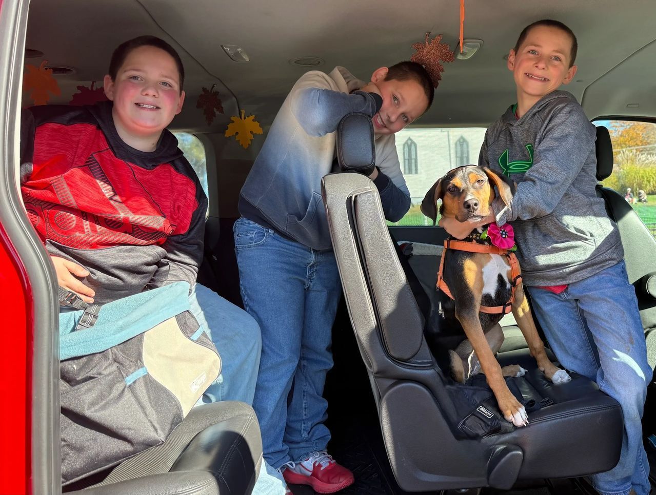 Three people and a dog smiling inside a car, with one person standing by a child car seat.