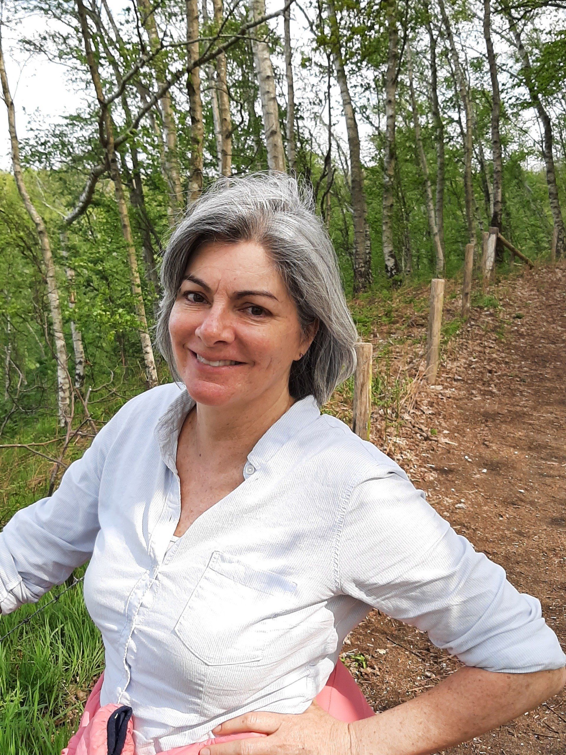 Woman with gray hair smiles in a wooded area, wearing a light blue shirt.