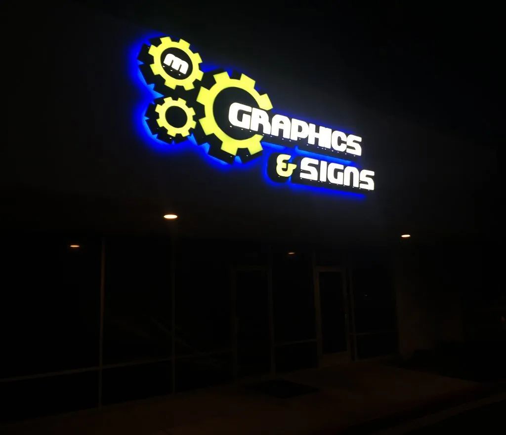 Neon gear-shaped sign reading “Graphics & Signs” glowing blue and yellow on a dark storefront at night