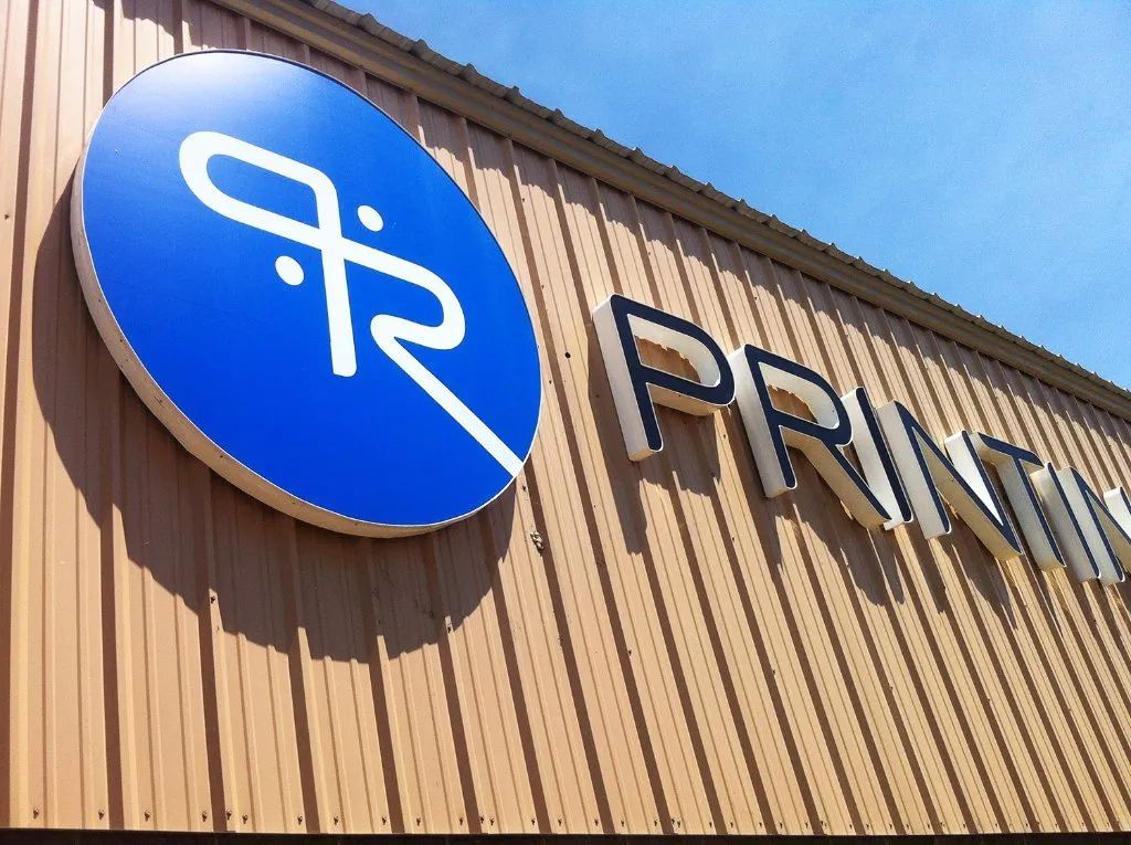 Blue circular sign with white logo on a tan building reading “PRINTI…”
