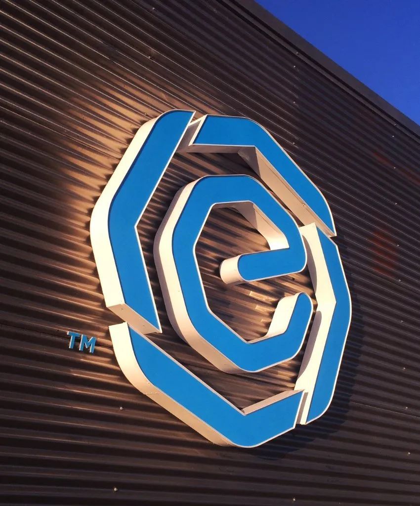 Blue and white geometric logo mounted on a dark corrugated wall at dusk