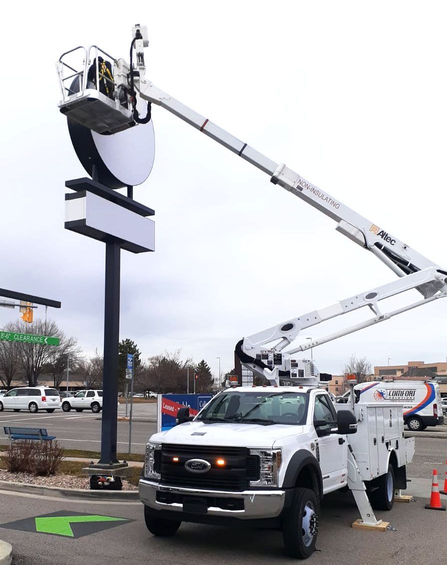 Two utility trucks with raised booms service a large roadside sign in a parking lot.