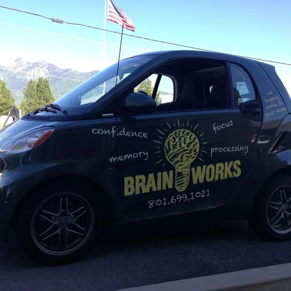 Black smart car with “BRAIN WORKS” logo and lightbulb graphic parked outdoors under an American flag.