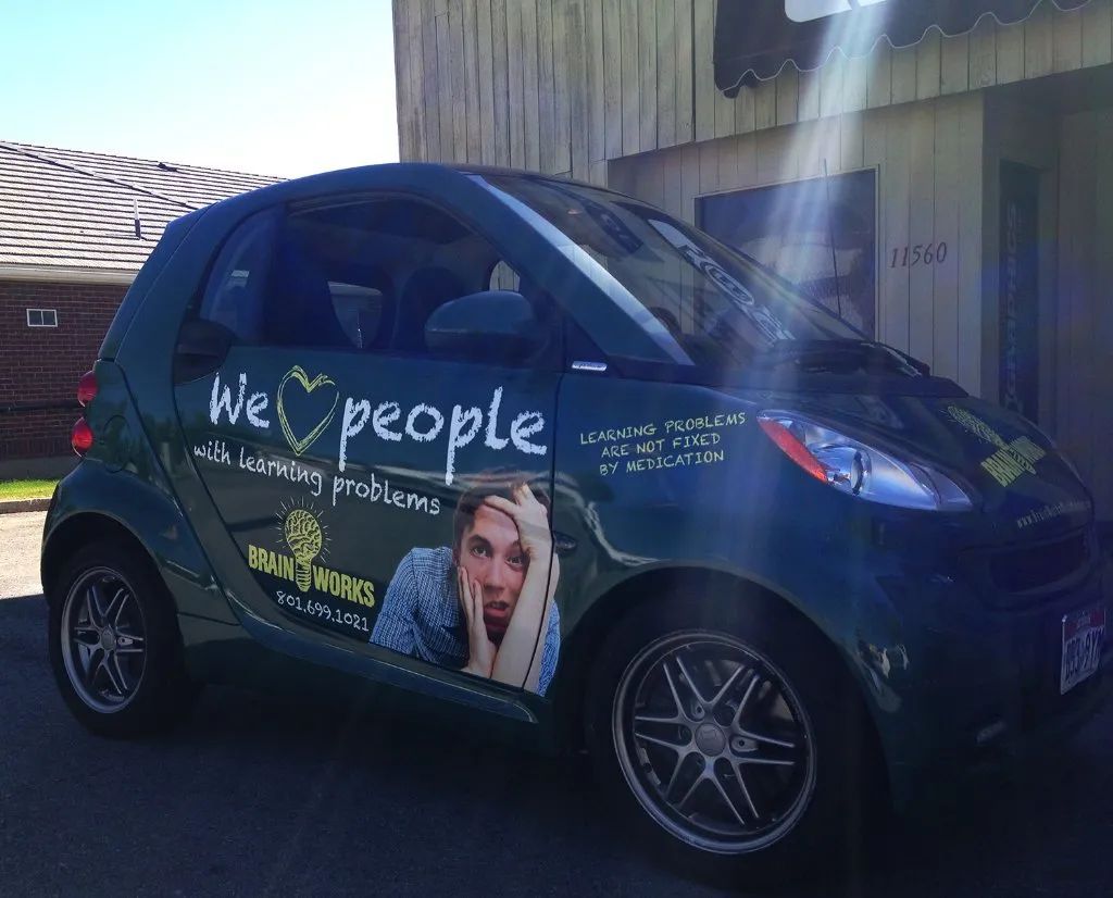Blue Smart car with “We ♥ people” promotional graphics parked outdoors in sunlight
