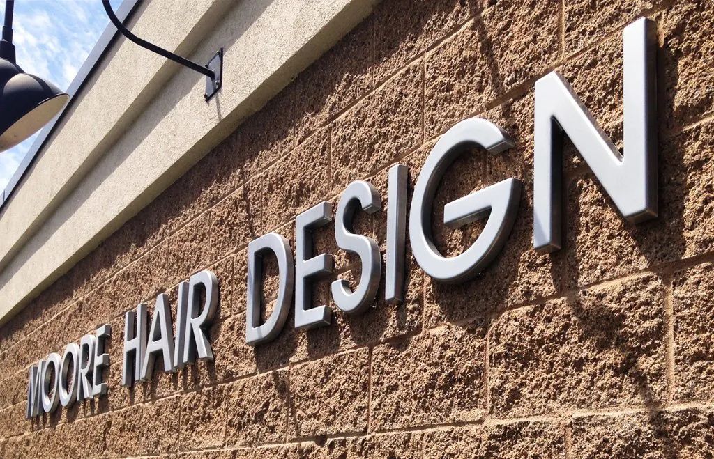 White “MOORE HAIR DESIGN” sign on a tan brick building exterior.