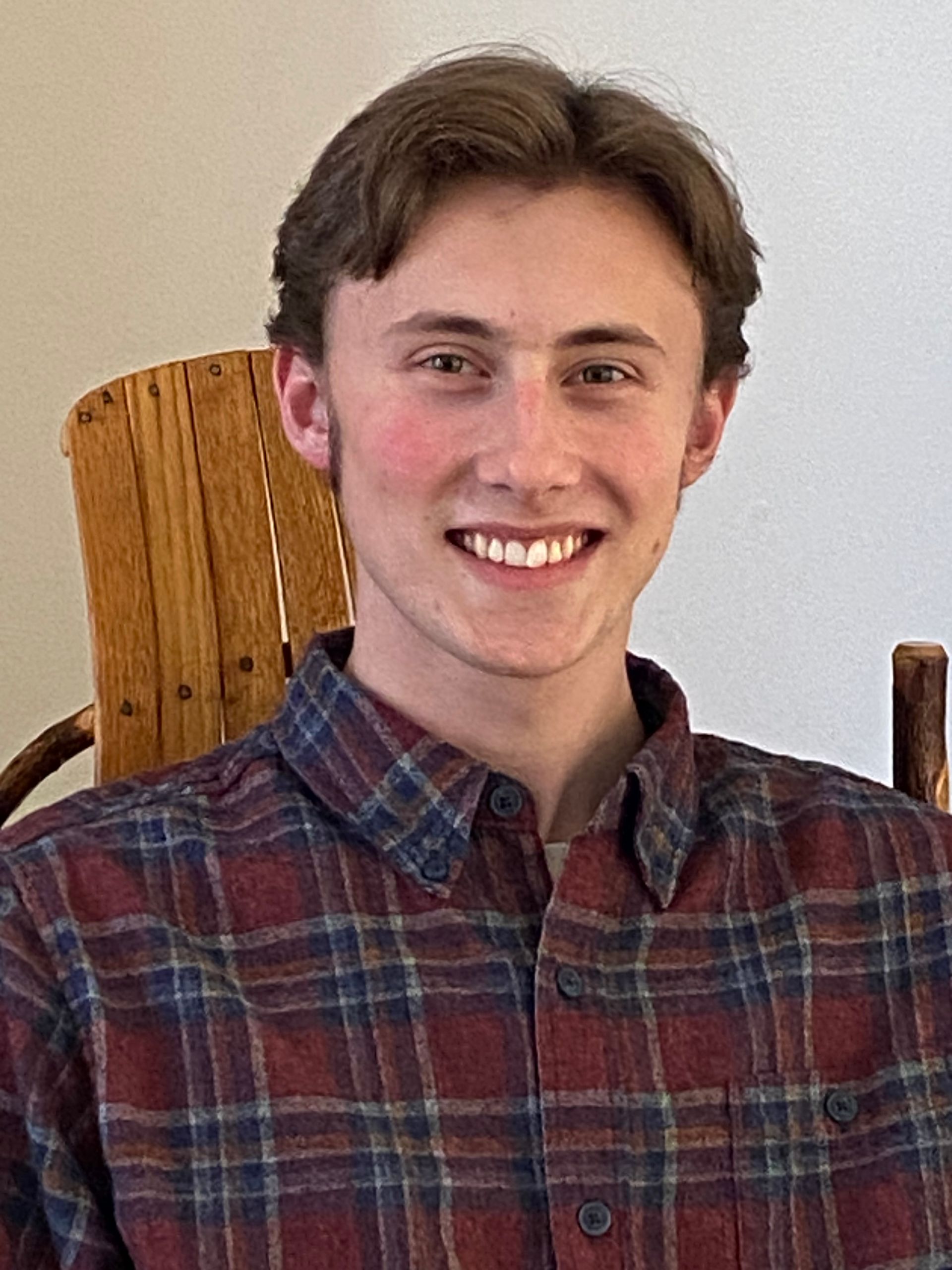 A young man in a plaid shirt is smiling while sitting in a wooden chair.