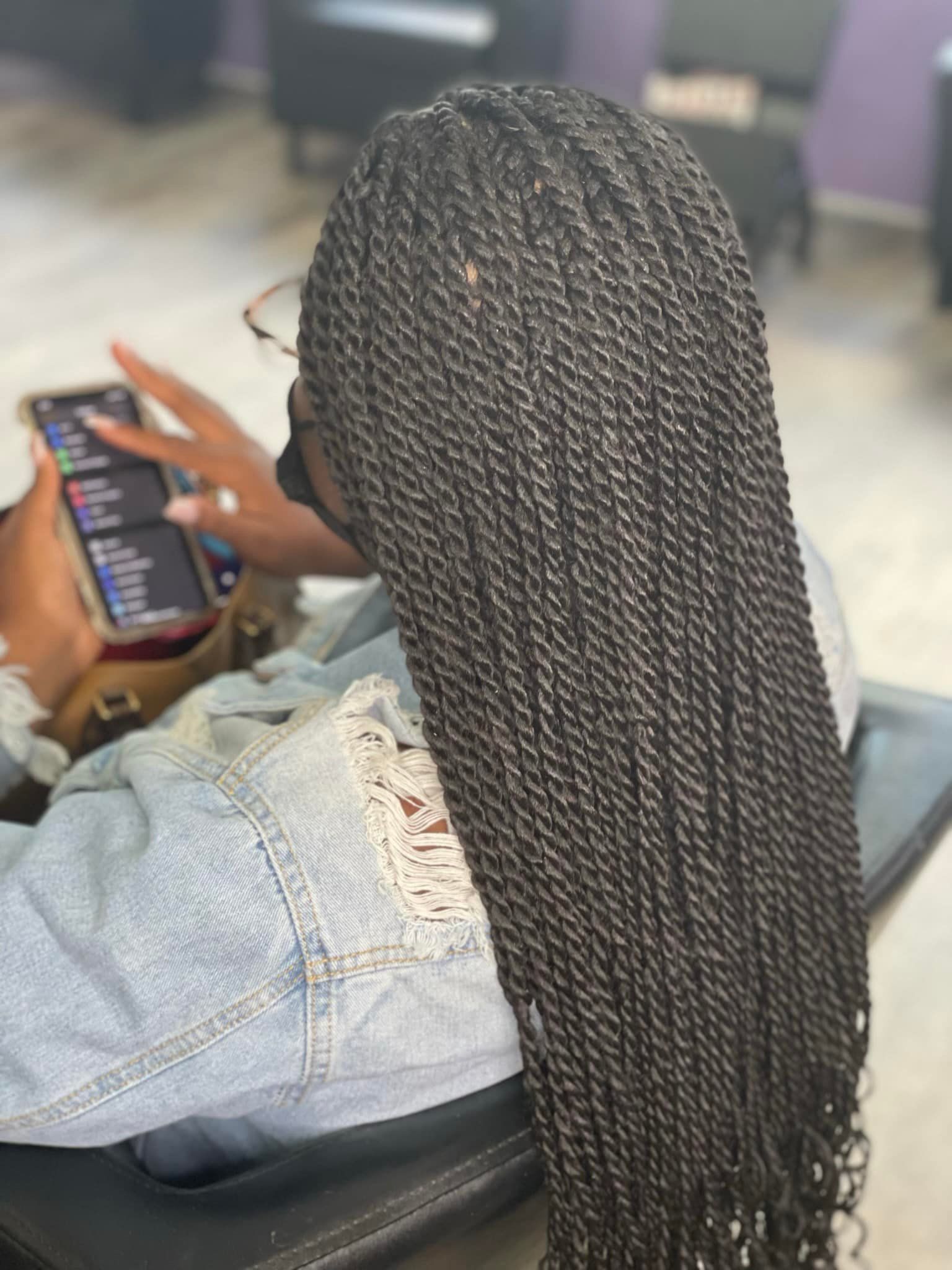 A person with long, dark, micro-twisted braids, seen from behind, holding a smartphone while seated in a salon chair.