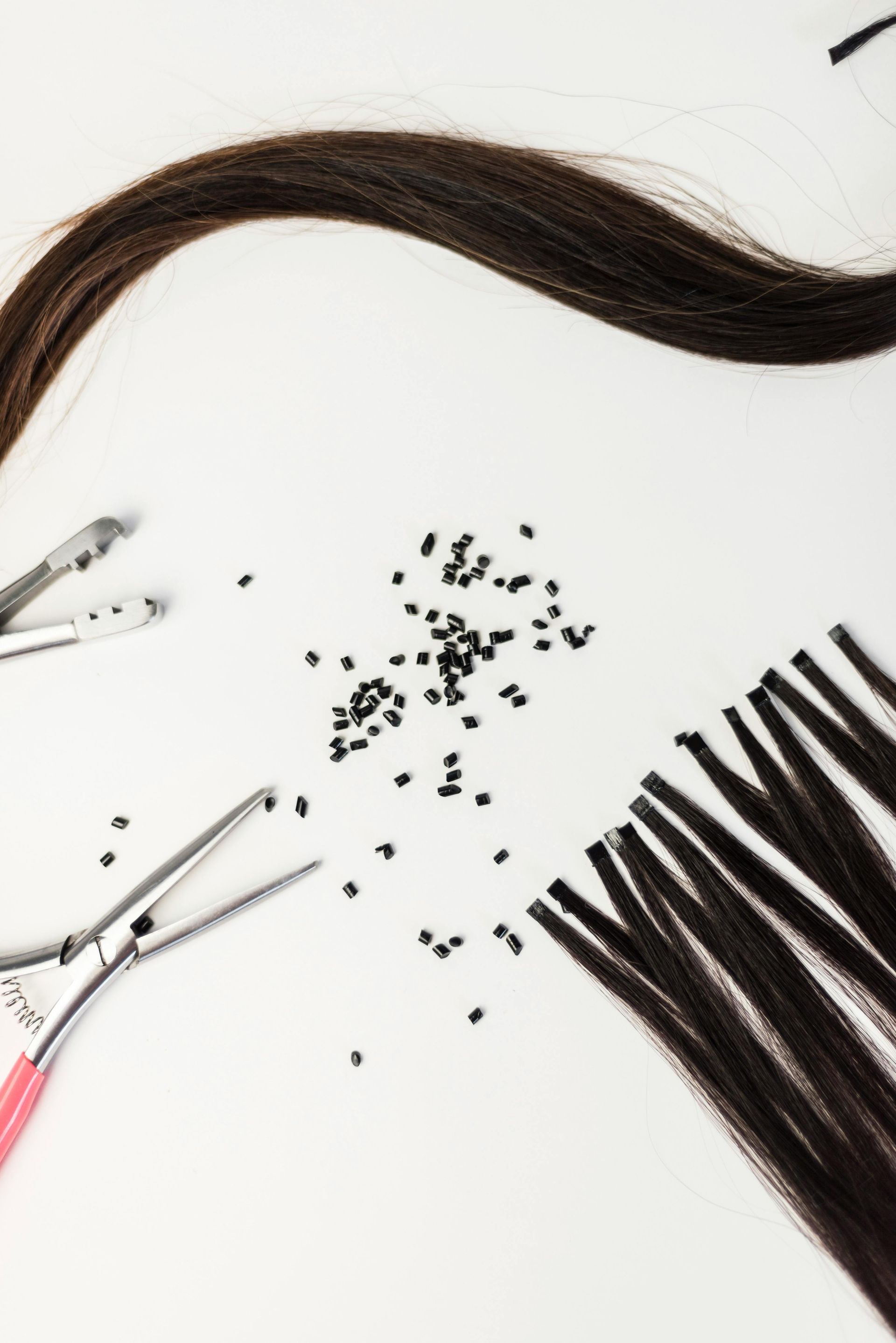 Hair extension tools, small black micro-rings, and brown hair strands arranged on a white surface.
