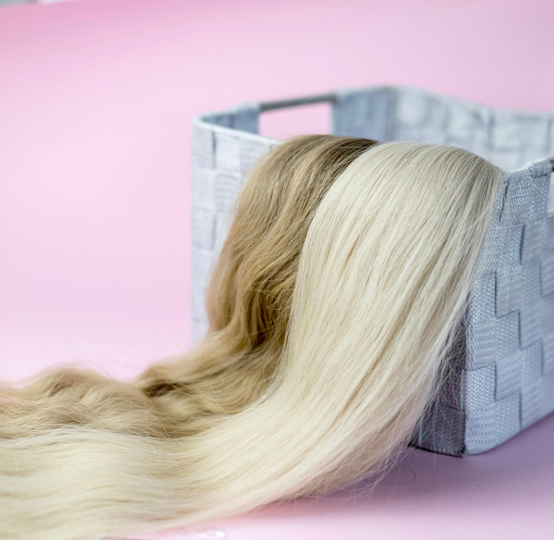 Two bundles of hair extensions, one blonde and one light brown, resting inside a gray storage basket on a pink surface.