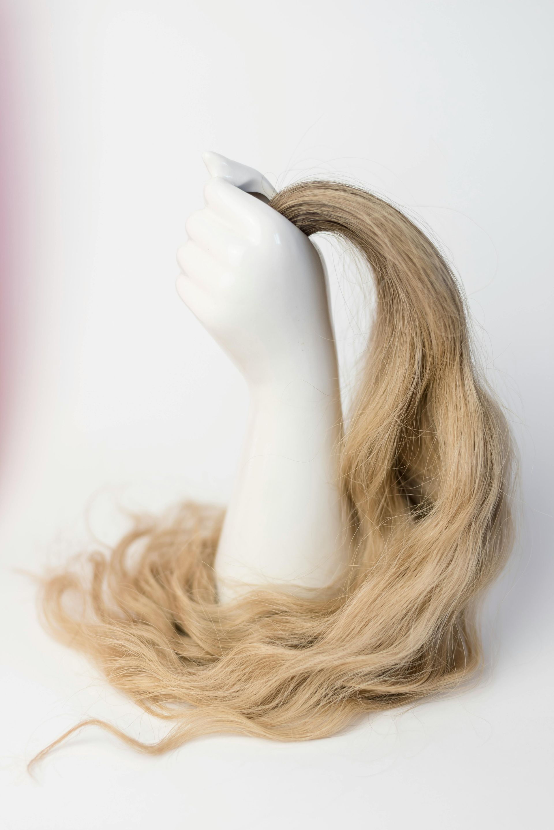 A white mannequin hand holding a long, wavy, light brown hair extension against a plain white background.