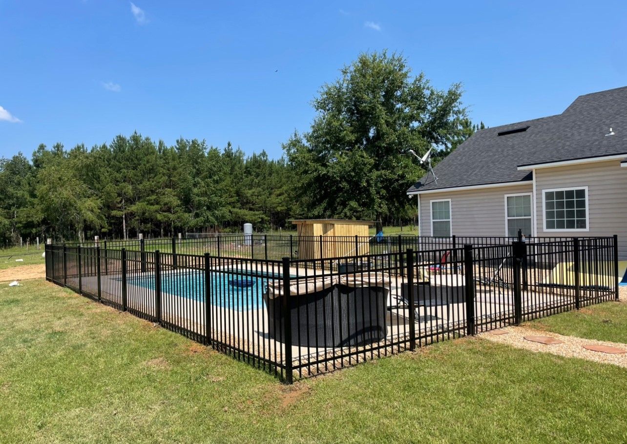 Black fenced-in backyard pool with patio furniture and a house in the background under a blue sky.
