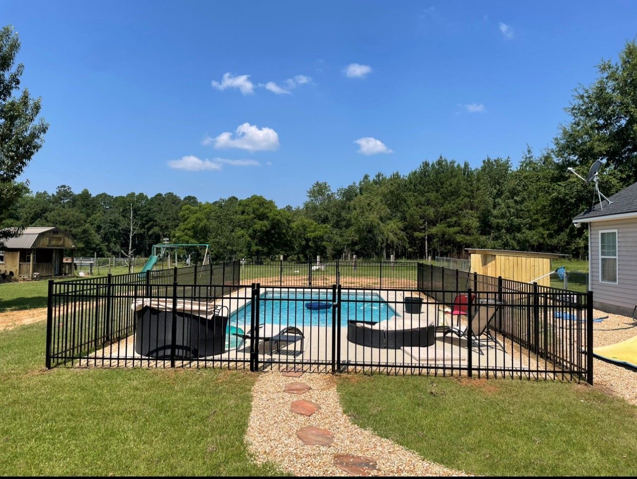 A backyard pool surrounded by a black fence on a sunny day. A stone path leads to the pool area.