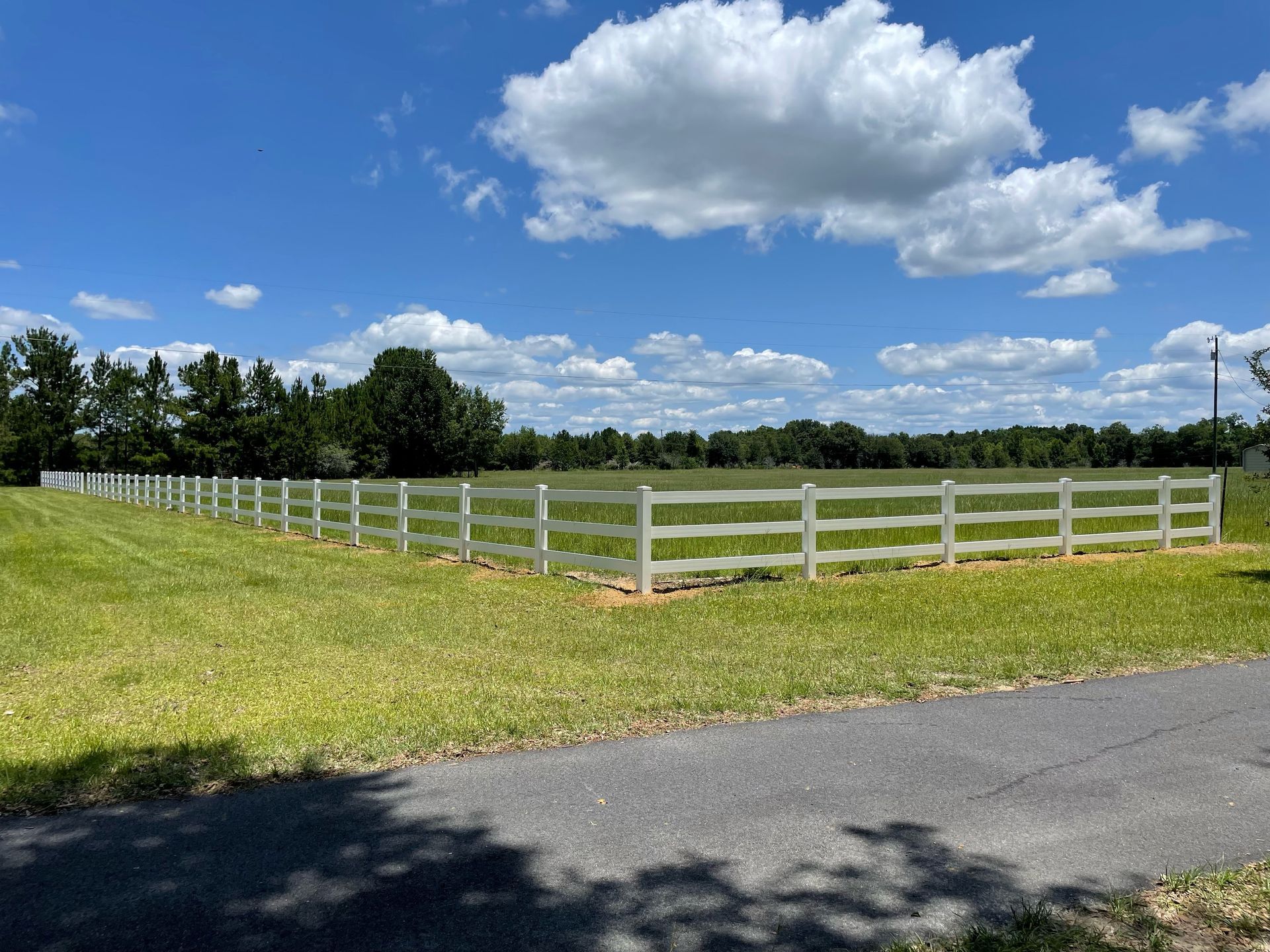 White fence encloses a grassy field under a blue sky with clouds; trees line the horizon.