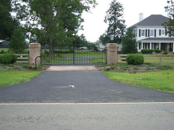 Black wrought iron gate with brick pillars, leading to a long asphalt driveway and white house.