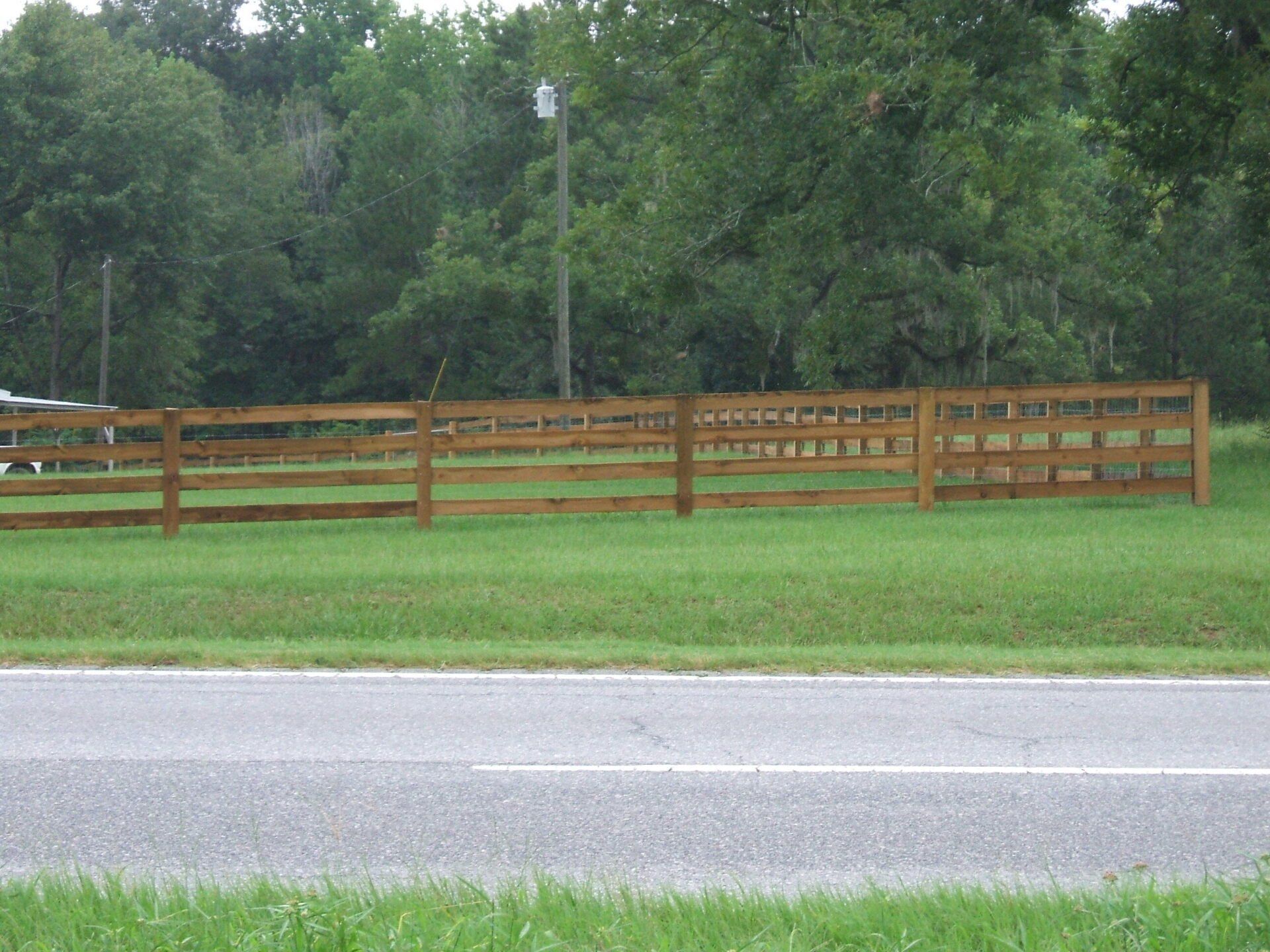 Brown wooden fence in a grassy field next to a road, trees in the background.