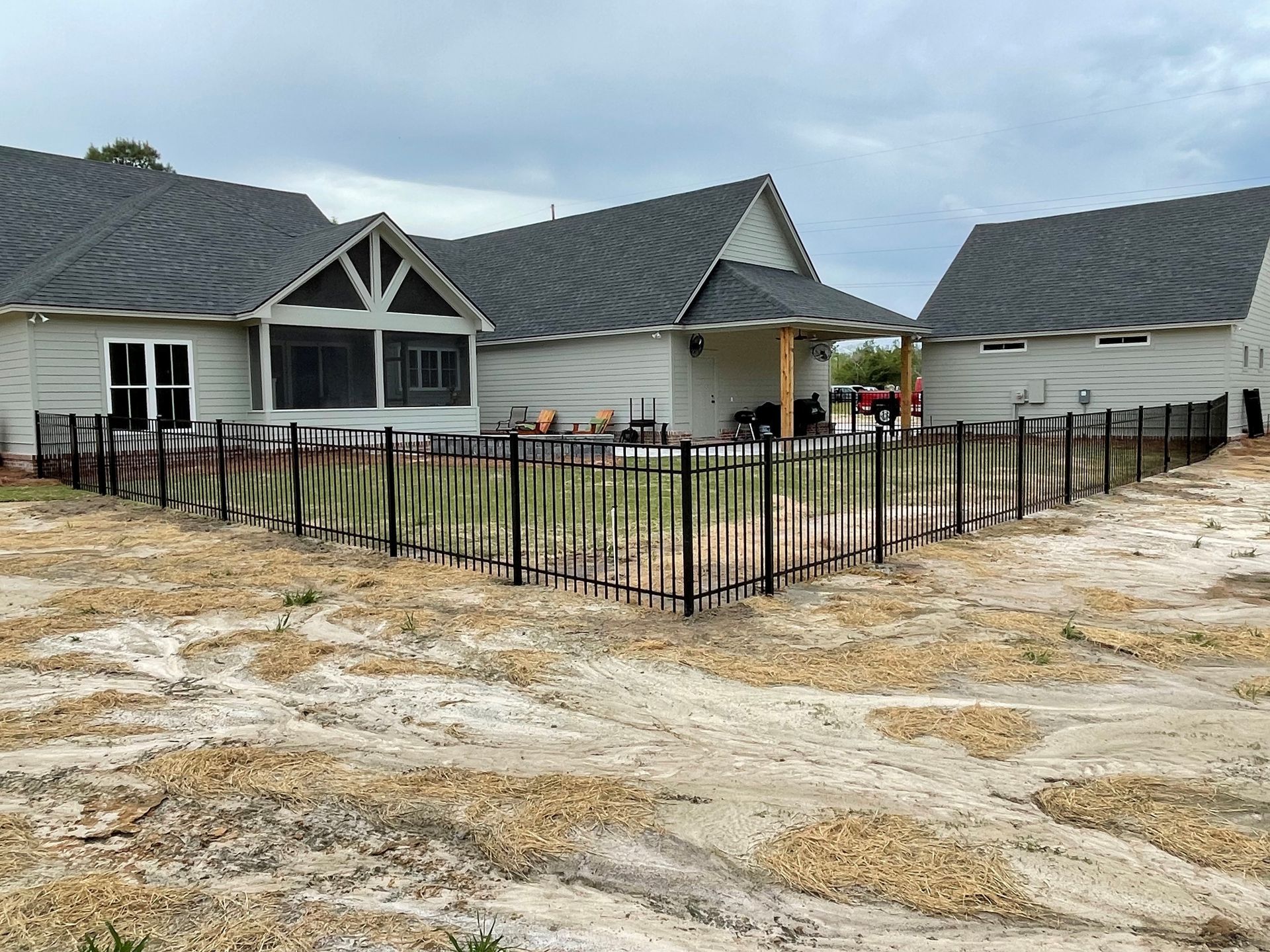 Black metal fence surrounds a grassy yard behind a light-colored house with dark roof under a cloudy sky.