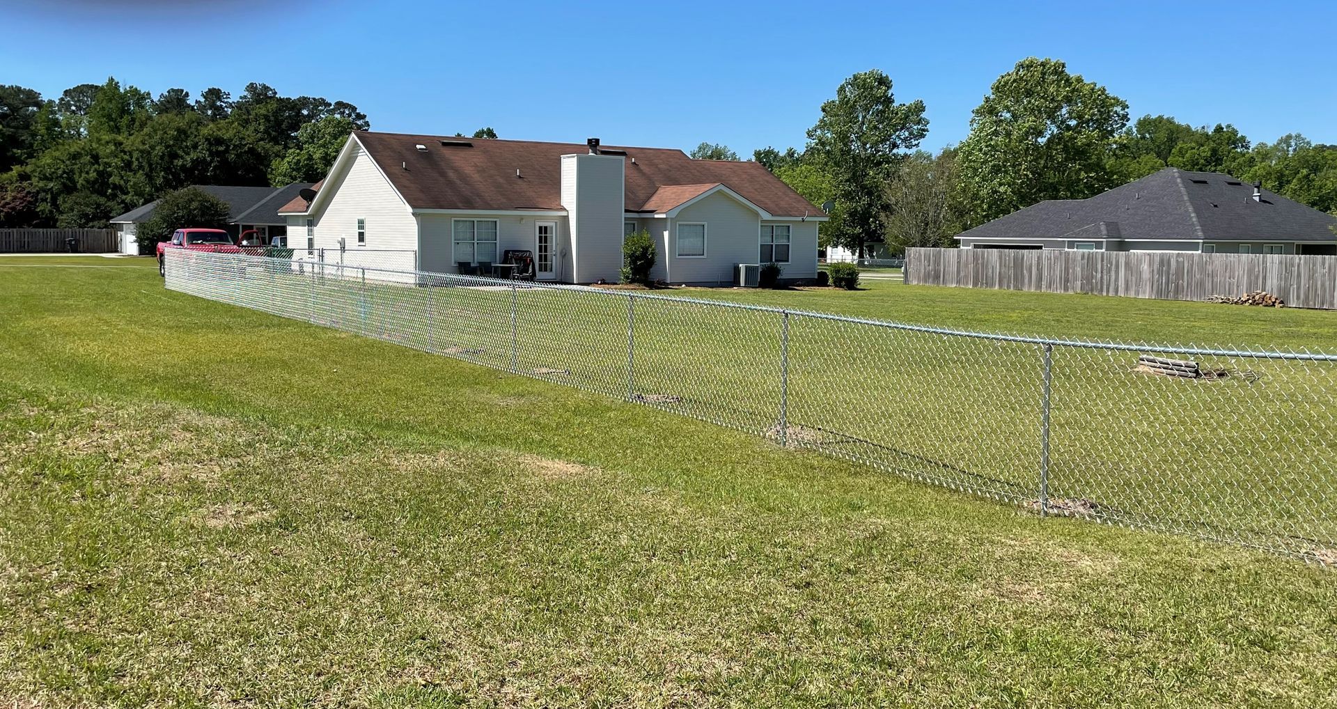 A chain-link fence surrounds a house with a green lawn under a blue sky, trees in the background.