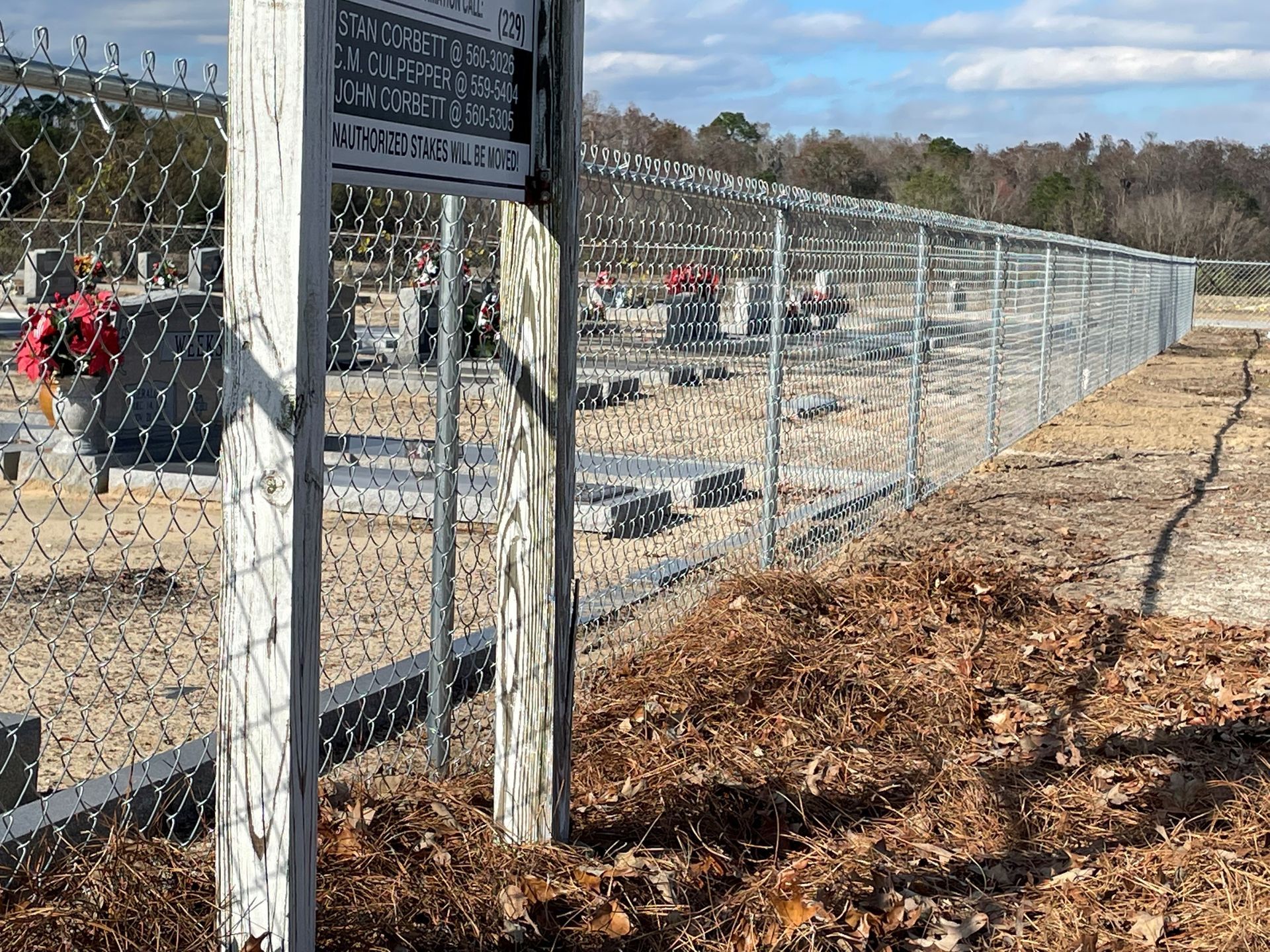 A cemetery with a chain-link fence. A sign is visible. The ground is covered with leaves.