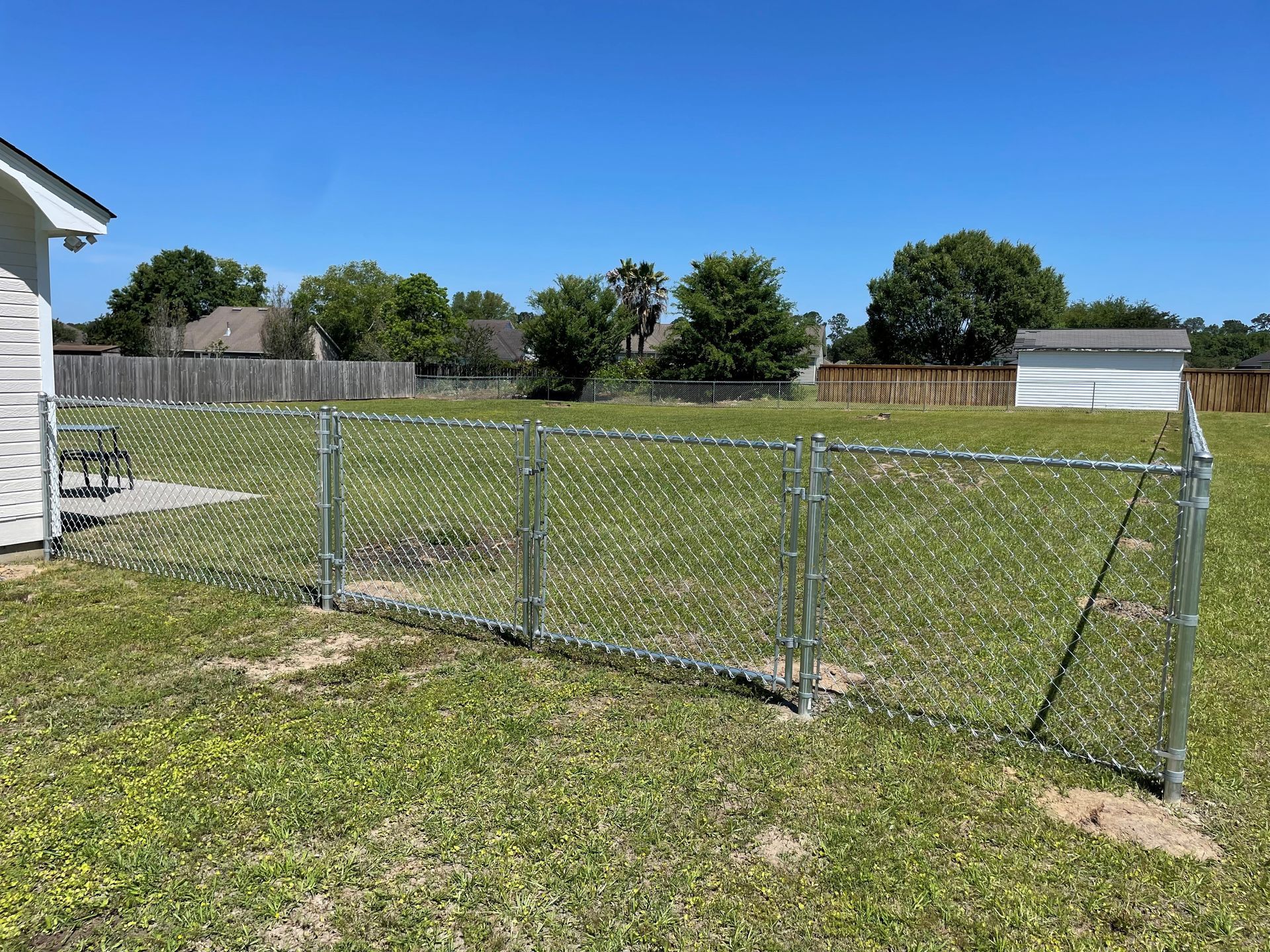 Chain-link fence in a grassy yard, partially surrounding a house with a blue sky background.