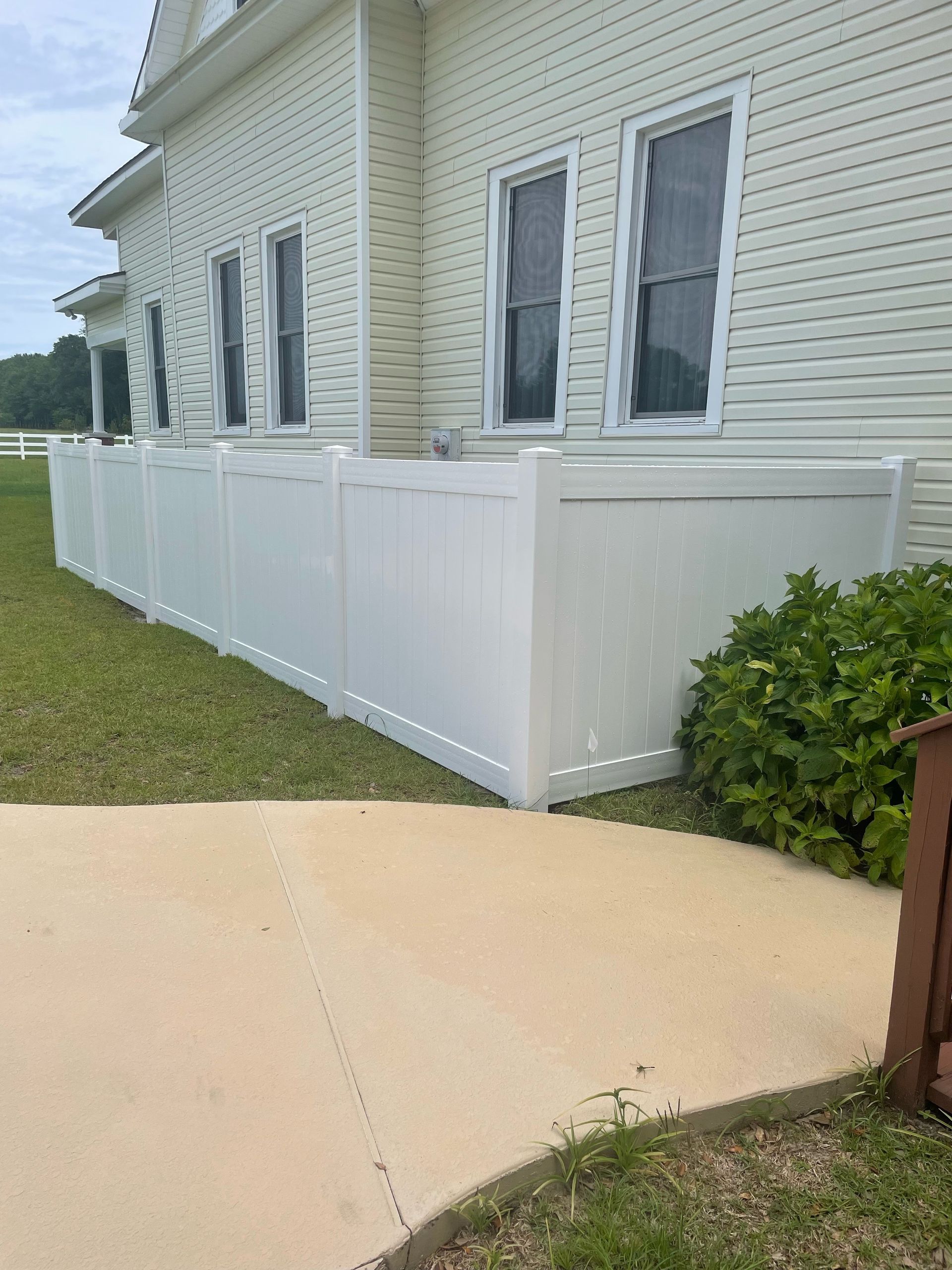 White fence along the side of a white building with windows, beside a concrete walkway and green grass.