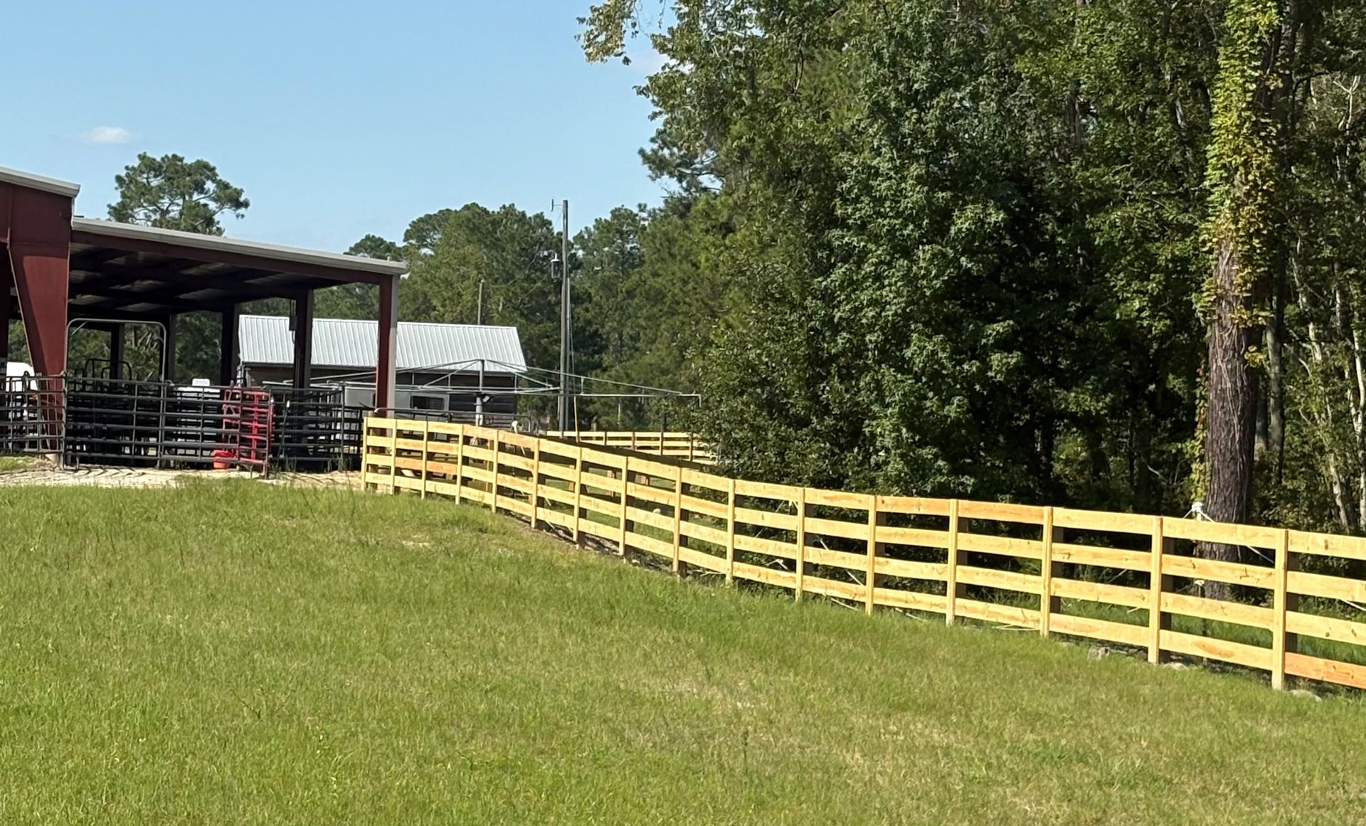 Wooden fence along green grass next to a red-roofed building. Trees in the background. Sunny day.