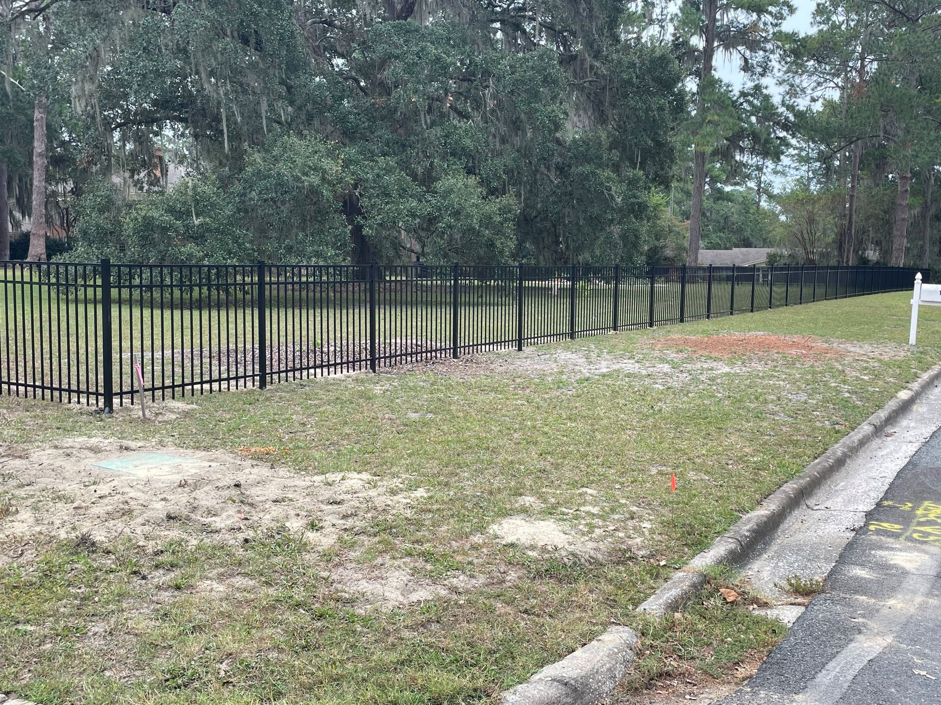 Black fence along grassy lot, trees in background, small sign on right.