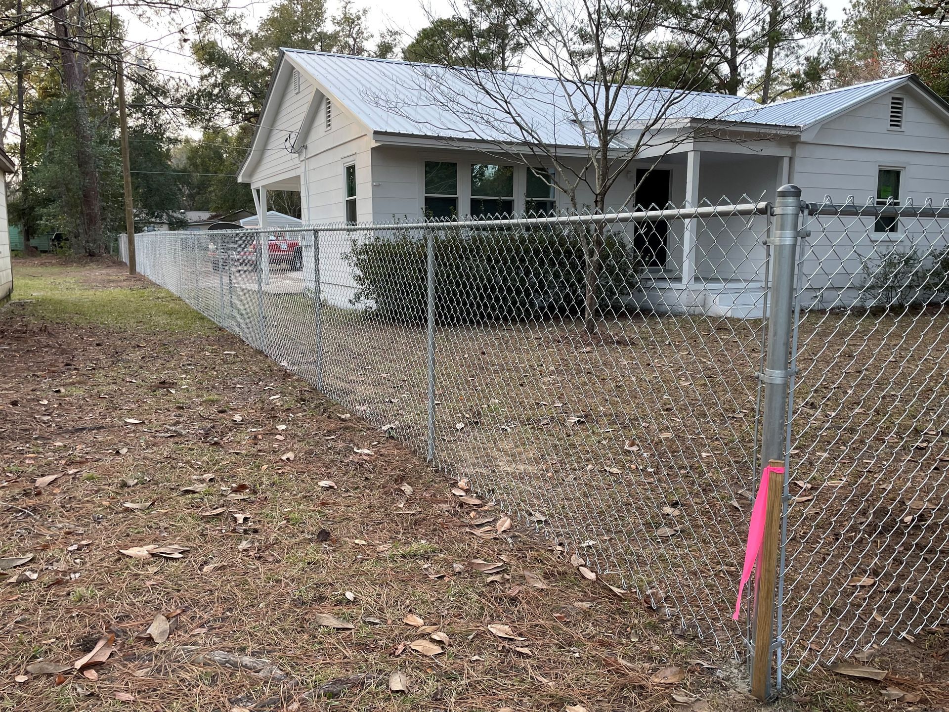 A chain-link fence surrounds a white house with a metal roof. A pink marker is on the fence post.