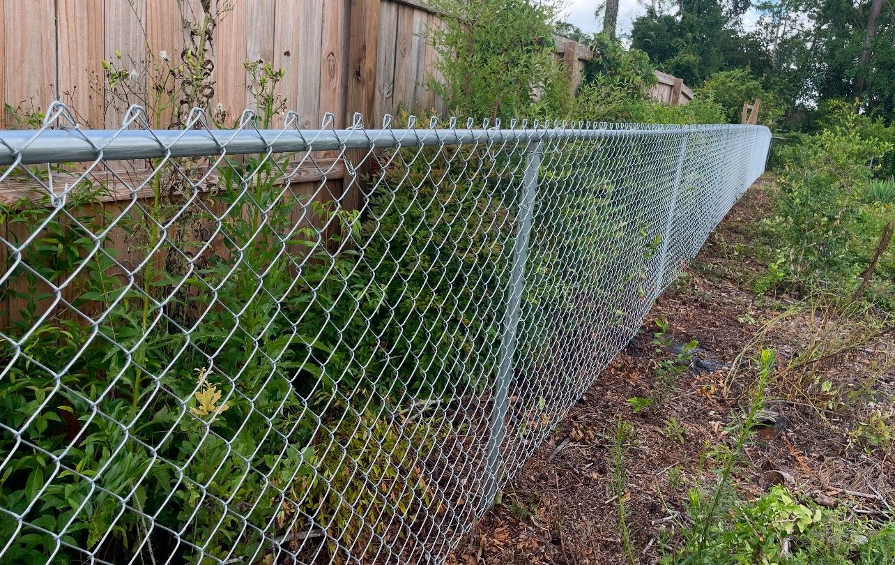 Chain-link fence running along a yard with overgrown vegetation, with a wooden fence in the background.