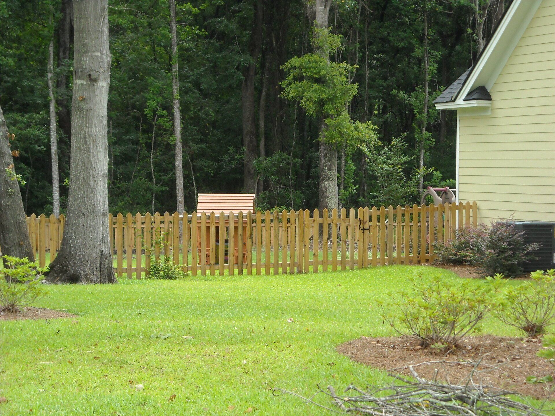 Lawn with picket fence, trees, and partial yellow house.
