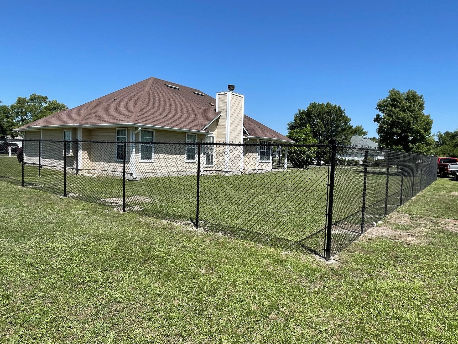 Black chain-link fence surrounds a grassy yard, beside a light brick house on a sunny day.