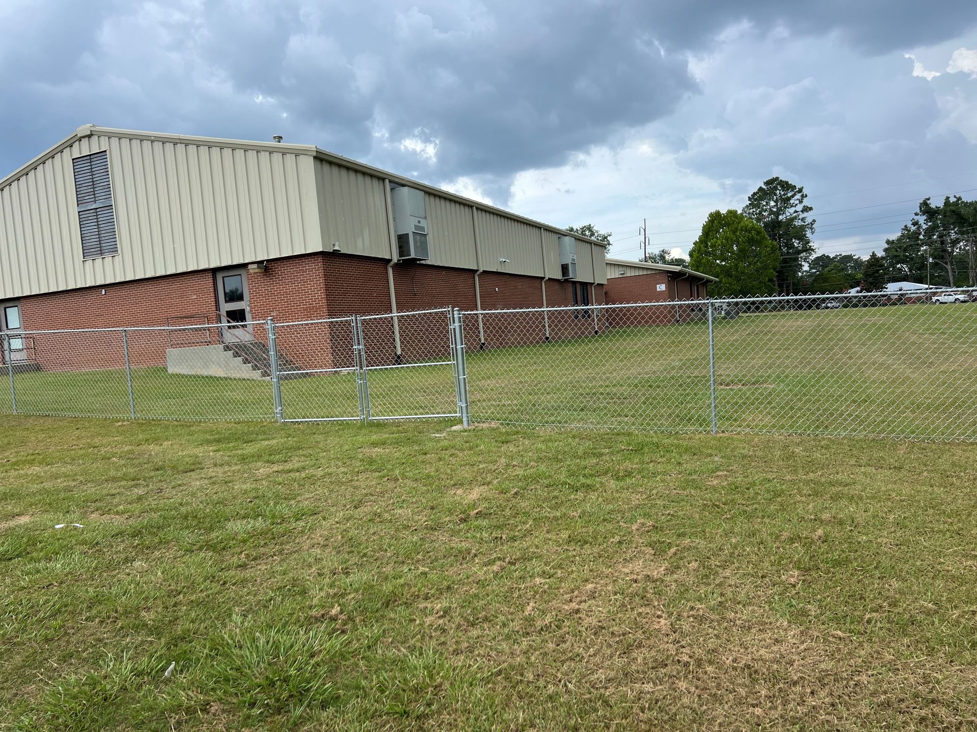 A low-rise brick and metal building with a chain-link fence on a grassy hill under a cloudy sky.
