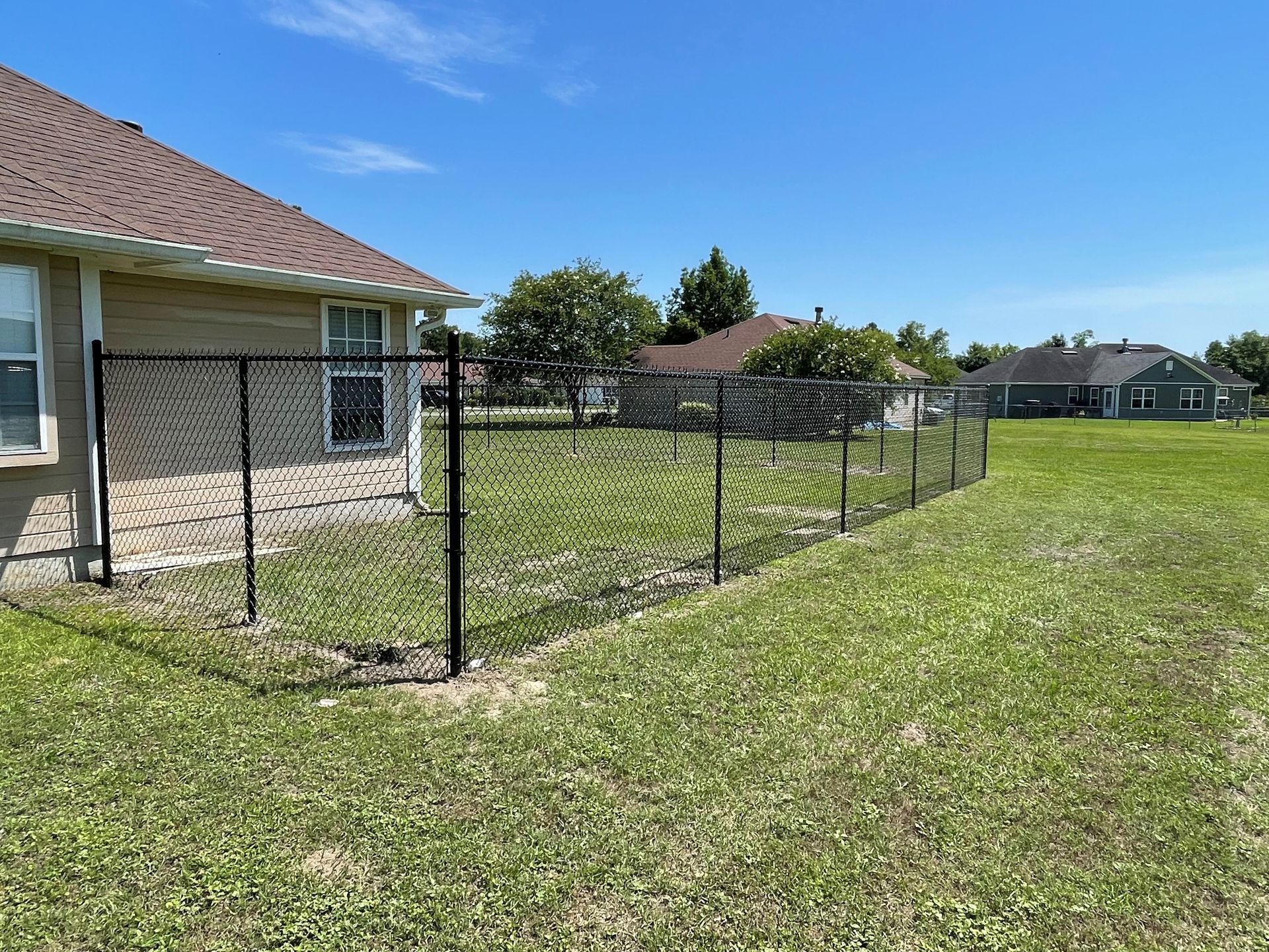 Black chain-link fence encloses a green backyard, with a house on one side and blue sky overhead.